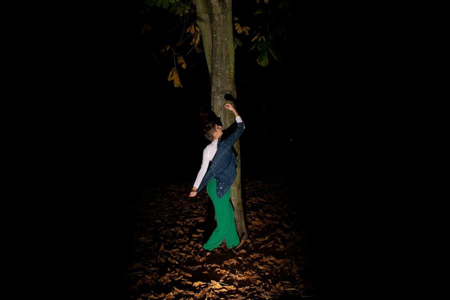 A woman wearing green trousers and a white top, with short blonde hair and fair skin, stands under a tree, with many orange leaves at her feet. She is mid-movement, with her right arm raised and her eyes looking towards it, taking off of a jean jacket.