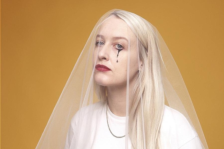 A girl with long blonde hair is looking at the camera. Her head is covered with a white veil. She has one painted black tear on her cheek. She is wearing a white t-shirt. The background of the picture is yellow.