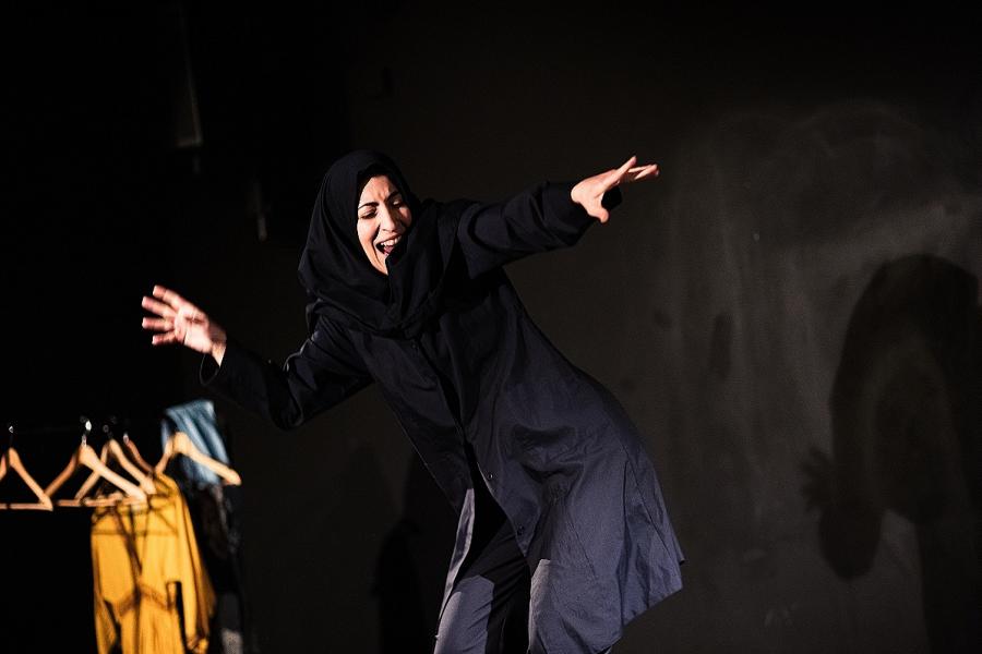 Sara, an Iranian women with light olive skin is dressed in a dark blue Iranian girls school uniform (blazer, trousers & headscarf). She smiles like a cheshire cat, dancing, with her arms out to her side. Behind is a bright yellow costume on a rail.