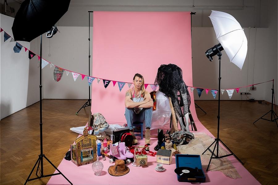 Man with long hair sat in a photographic studio in front of a pink screen and bunting.