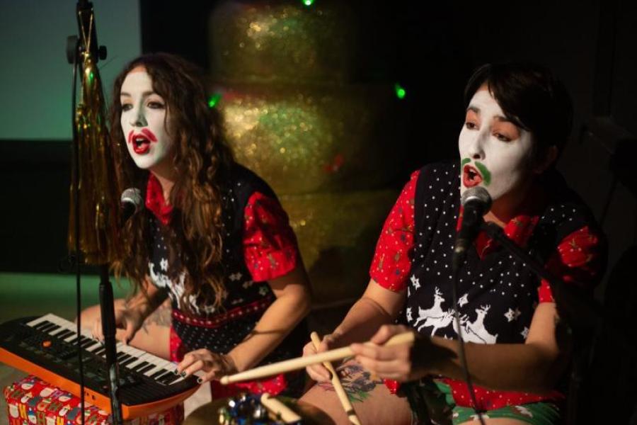 Two performers in a white facepaint with moustaches drawn on, playing keyboards and drums. They're wearing christmas vests over christmas shirts. Because why not?