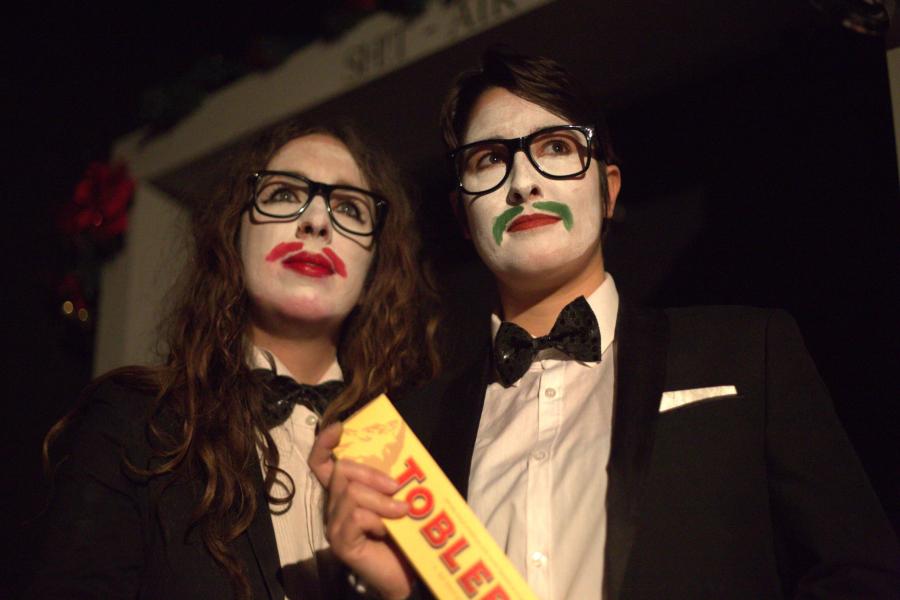 Two performers in a white facepaint with moustaches drawn on. They're wearing tuxedos and holding a giant Toblerone. Because it's Christmas.