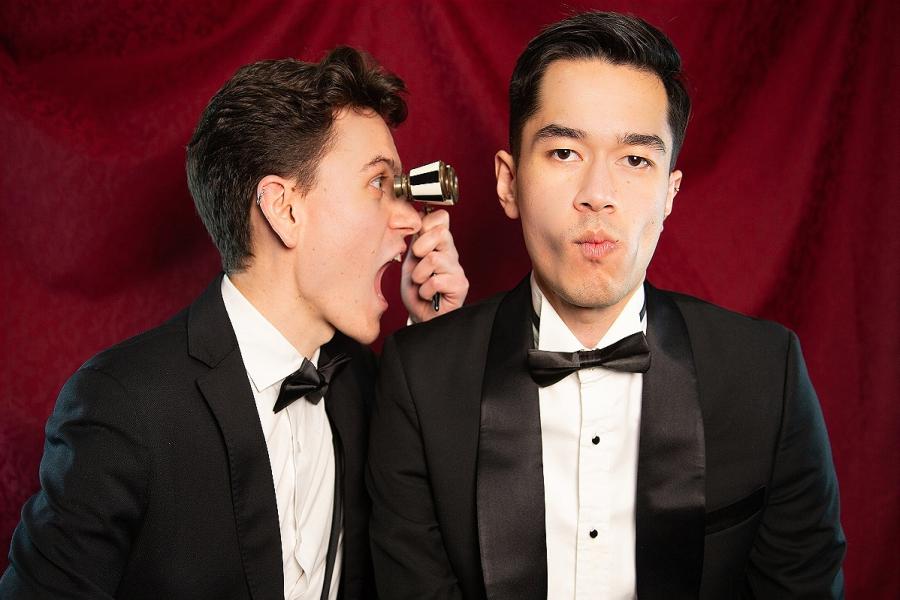 Gabriele and Joey in suits in front of a red drape. Gabriele looks at Joey through black and white opera glasses, while Joey looks at the camera and purses his lips in disapproval.