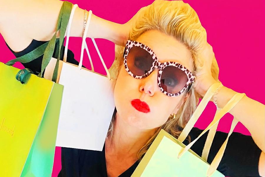 Woman wearing leopard-print sunglasses and red lipstick poses against a bright pink background, holding multiple shopping bags, swamped by consumer goods—playful yet overwhelmed.