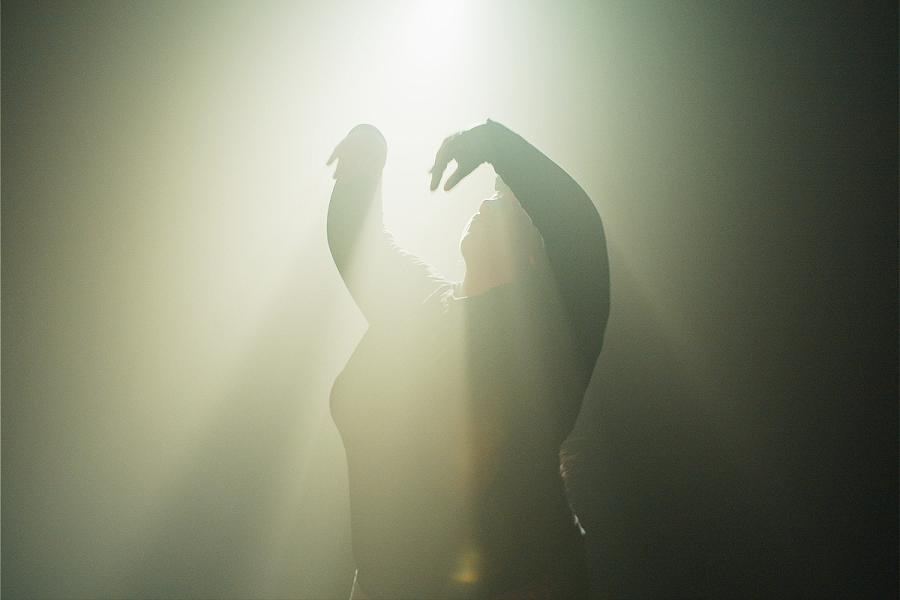 A young woman dancing in a soft pool of hazy light. Her arms are up, her eyes closed, her head back. She lets the light bathe her from above as, her raised arms form shadows across her body.