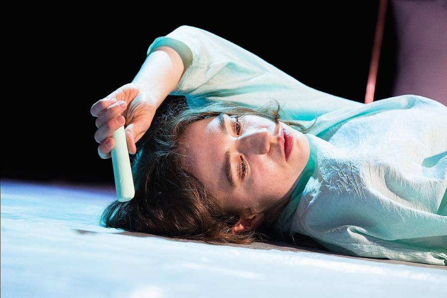 A close up photograph of a white, brunette, afab performer lying on the floor, against a dark background. They are reaching their left arm over their head, holding a piece of light blue chalk. Their eyes are downcast and they are in a pale blue shirt.