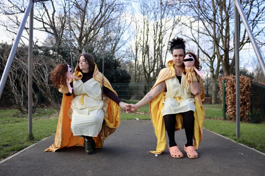 Photo of two people sat on a swing set. They are both wearing white togas with gold capes and are each holding identical felt puppets who are wearing gold and have long brown hair. The person on the left is white, has wavy brown hair and is gazing into th