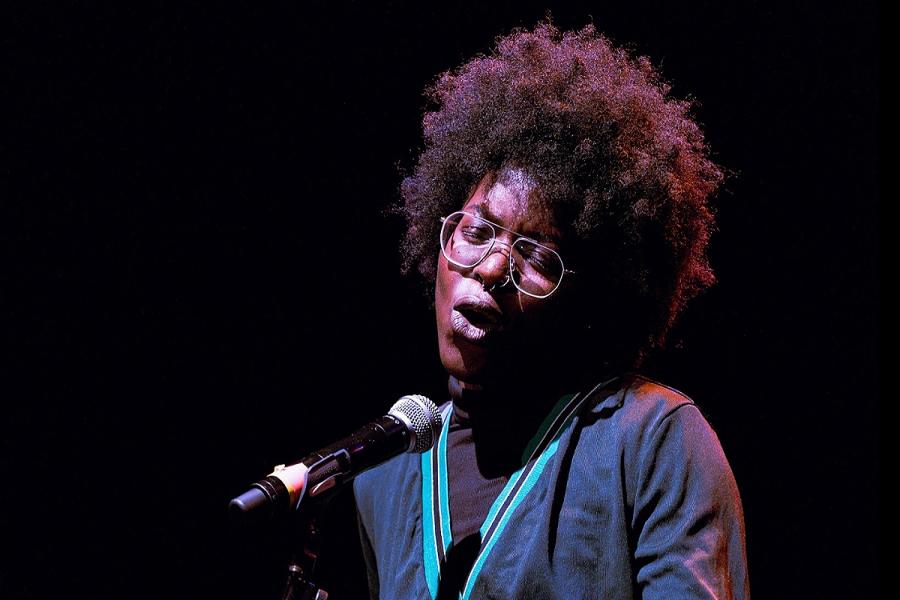 Princess Bestman (Black person with afro and glasses) standing on stage against a black background. They are mid performance speaking into into a standing mic.