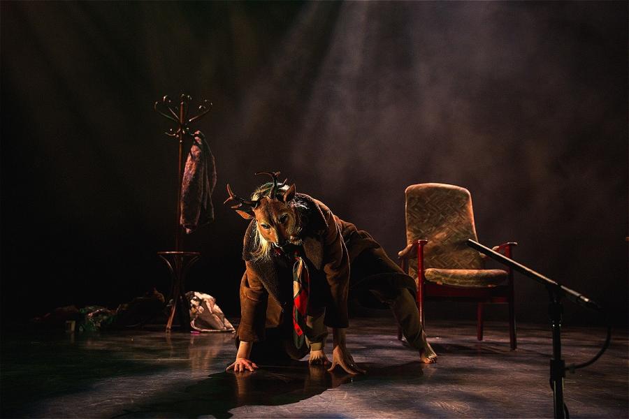 Portrait - A woman in a deer mask sits in a chair Landscape - A woman stands on all fours wearing a deer mask and big brown coat