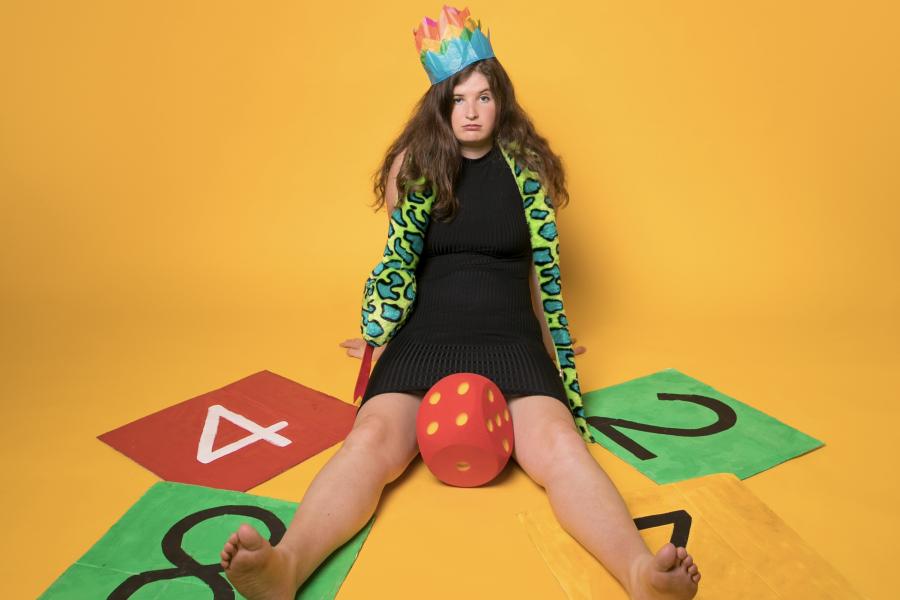 Show poster: A woman sits on top of giant colourful numbers with a big dice between her legs and wearing 3 Christmas cracker crowns.
