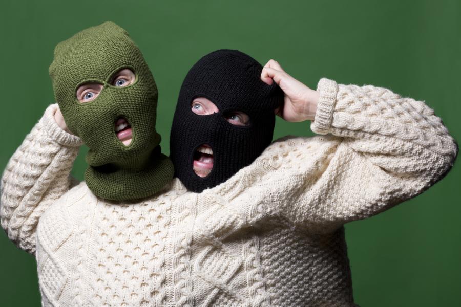 Two figures, wearing balaclavas, and squashed into a large cream, intricately patterned wool jumper hold their hands up, indicating a possibly alarming situation. The figure on the left wears an olive green balaclava, the figure on the right wears a black