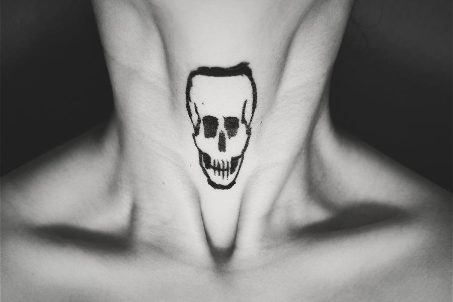 A black and white photograph of someone's neck, from the collarbone to the chin; with a skeleton skull drawn on the surface of the throat area, in black marker