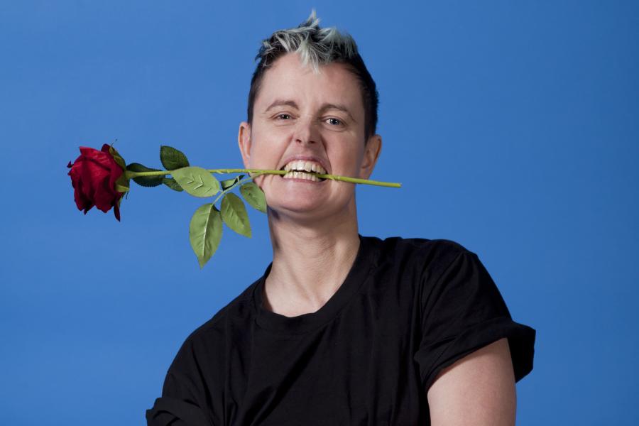 Photograph shows Am Stubberfield (slim white trans masuline person with short blue hair wearing black t shirt) Am has their arms crossed and grips a single red rose between their teeth with a growling sexy expression on their face