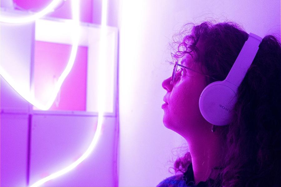 A woman with glasses and curly brown hair wears white headphones and looks up towards a white box with purple lights hanging from it.