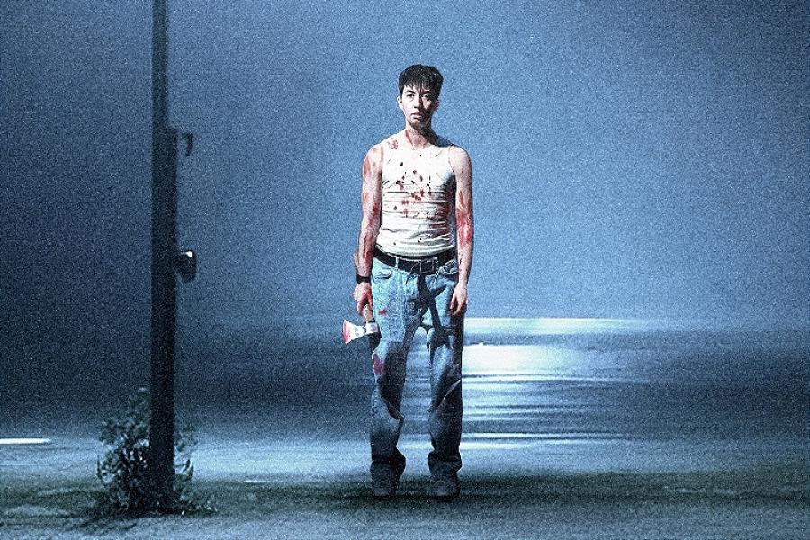 A short, white masculine presenting person with short brown hair stands in the light of a street light wearing a white vest and blue jeans spattered with blood. They are holding an axe with blood on the blade and staring vacantly in to the camera.