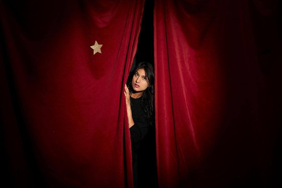 A dark haired woman peers out from a red velvet stage curtain and points at a star which she is desperate to reach.