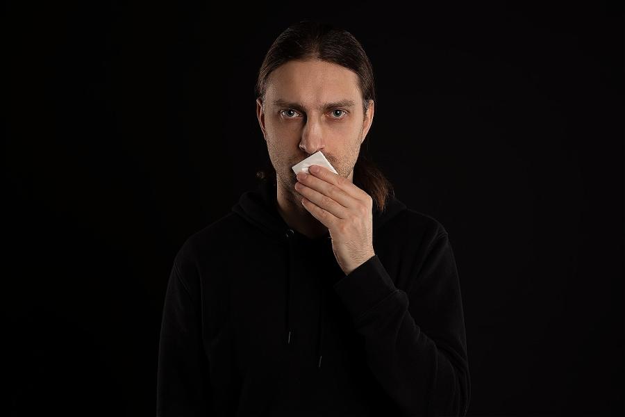 The head of a young man on a black backdrop covering his mouth with a handkerchief.