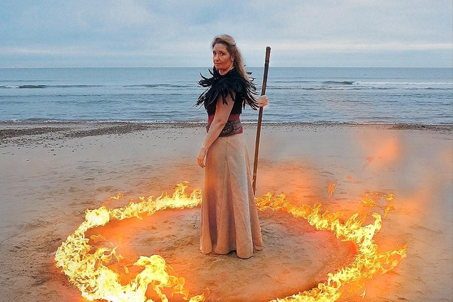 A woman stands on a beach. Around her in the sand there is a circle of flames. The ocean is the background. She is holding a large stick/staff looking into the camera.