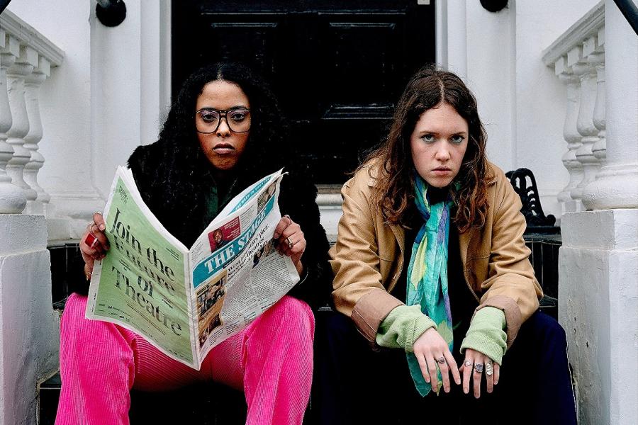 Two young female presenting people sat on the steps outside a house. One is wearing a black fur coat and pink trousers and glasses and is holding a copy of The Stage. The other is wearing a brown leather jacket, a blue scarf and navy trousers.