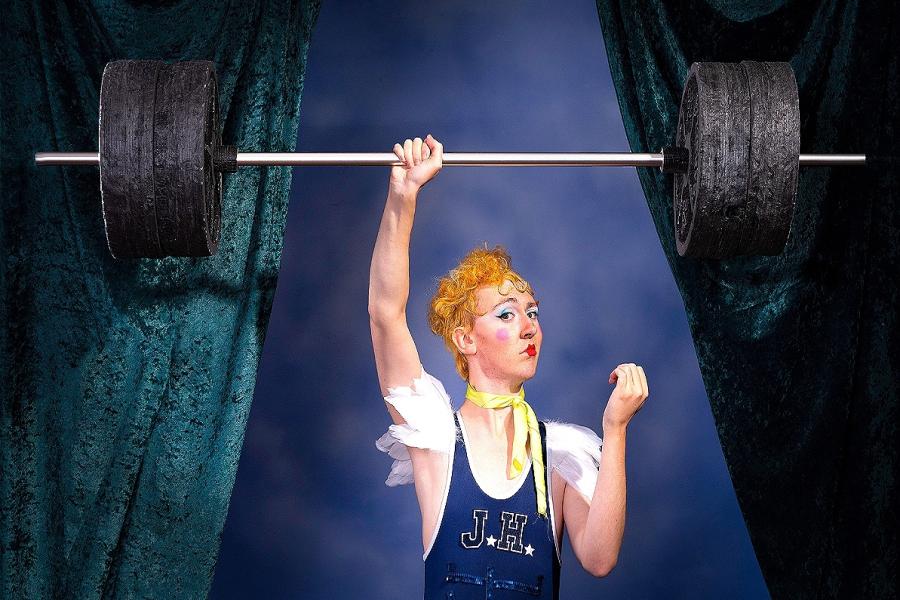 Lachlan poses as Jack Hammer, a circus strongman - with curled blonde hair in a 1920s style, wearing a blue singlet with white feather shoulders and blue sequined abs, he lifts a huge barbell with weights above his head with one hand.