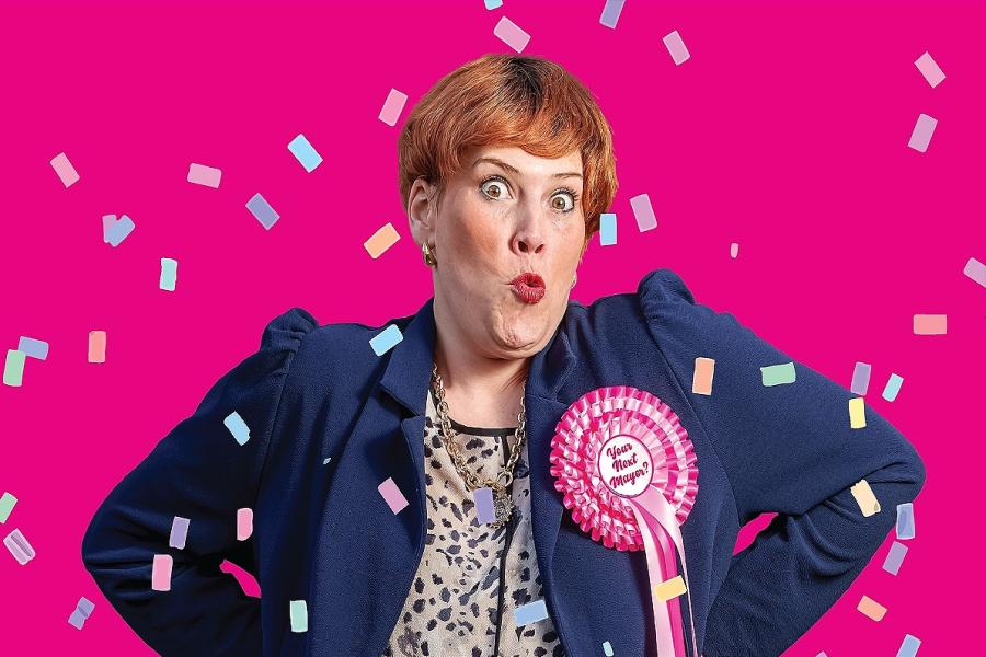 A woman in a blue suit with a playful surprised expression against a bright pink background, with colourful confetti around her. She wears a large pink rosette reading “Your Next Mayor?”.