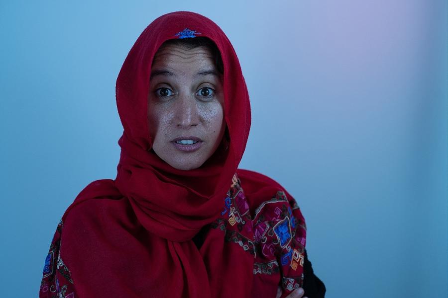 A woman with a red head covering stares directly into the camera lense infront of a blue background with a light gradient.