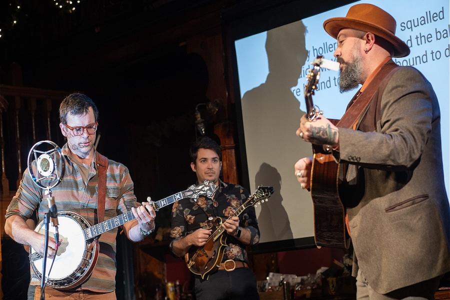 Banjo, guitar, and mandolin player on stage, in front of a projector showing lyrics of a song about President William McKinley