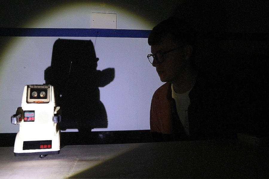 A toy robot in spotlight, its shadow cast across the wall behind it. To its right, a human figure (wearing glasses) staring at it in silouhette/shadow.