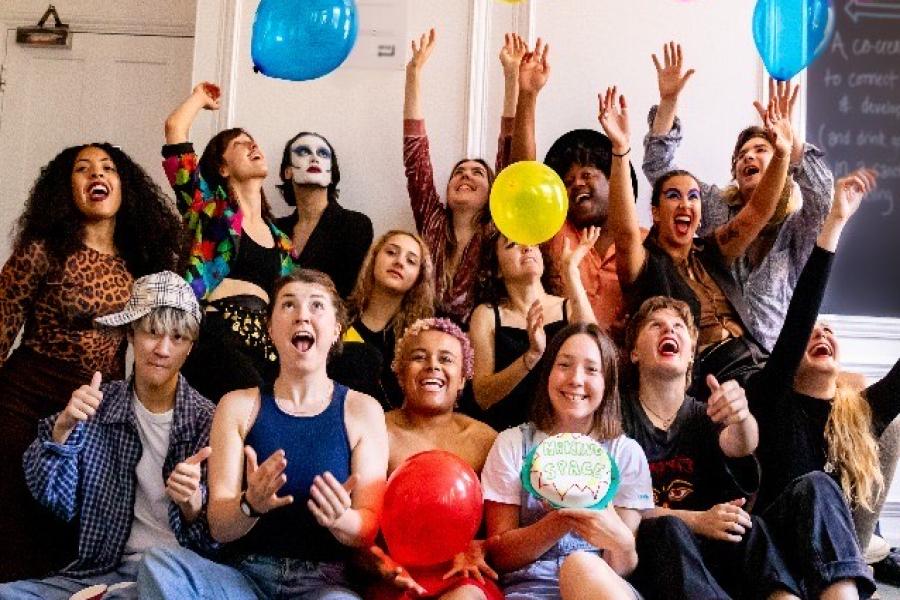 14 young adult theatre makers sit on a sofa with balloons around them. They wear colourful clothes. Some wear face paint, some smile, some pout, some hold a cake or cup. The feeling is one of expression, joy and a mix of wonderful personalities. 