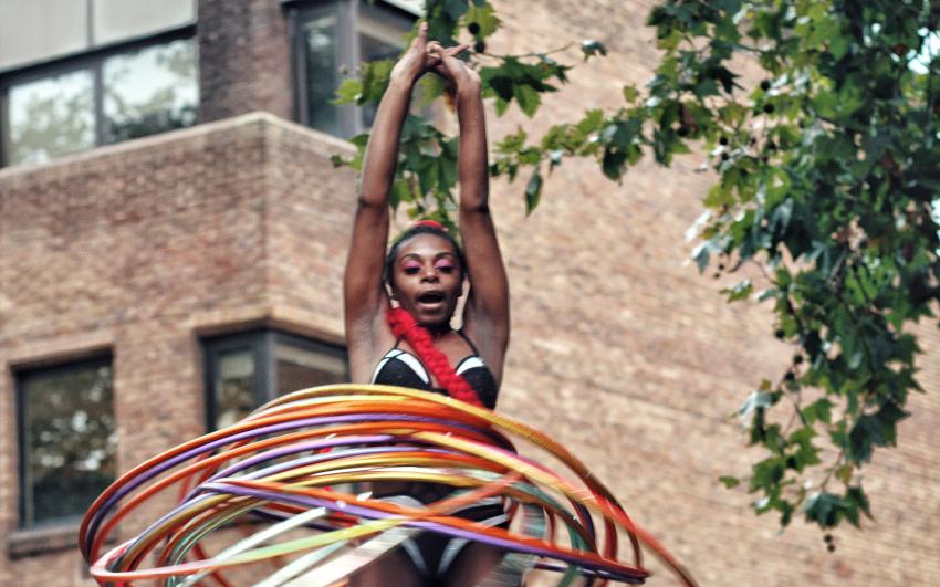 Photograph of a performer with lots of colourful hula hoops in a black leotard performing outdoors in Tolmer's Square.