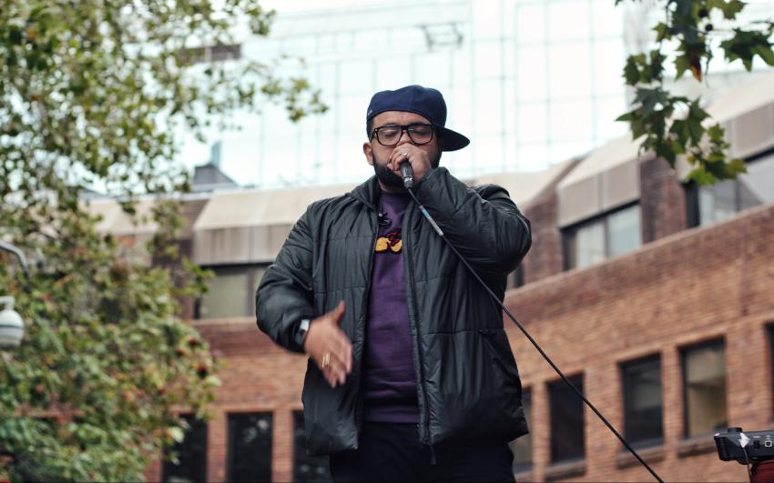A performer in glasses and a sideways cap speaking into a microphone in Tolmer's Square.