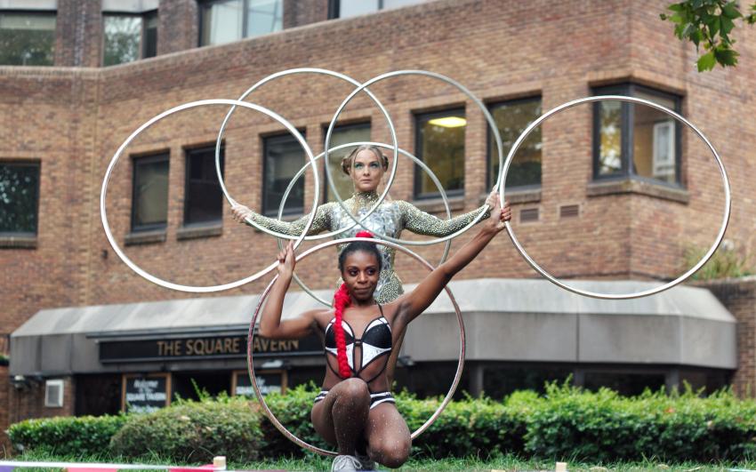 Two people wearing dance leotards, both holding multiple silver hoolahoops. One person is crouching in front of the other, who is standing.