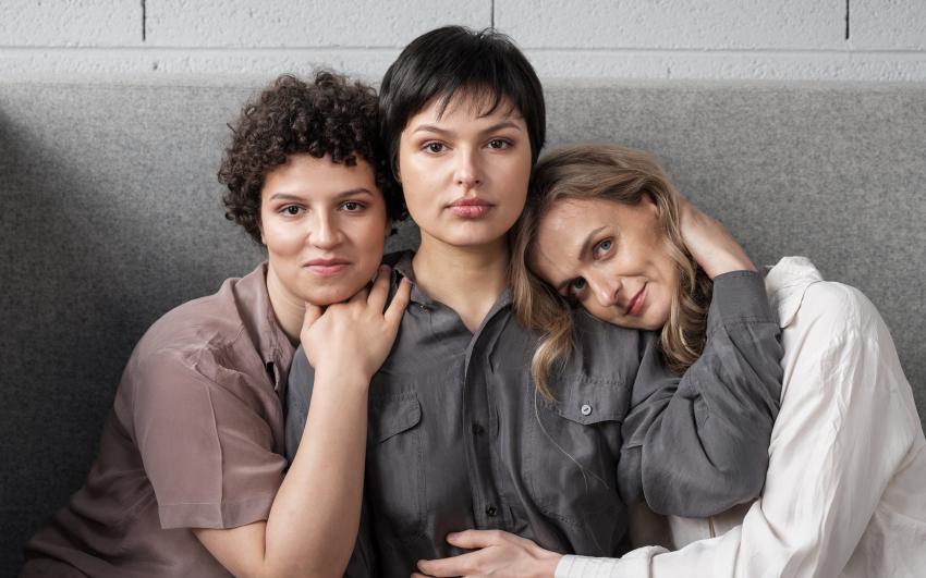 Three women of different ethnicities sat on a grey sofa, seen from the waist up. A white brick background. The women are looking straight at us. They are confident.