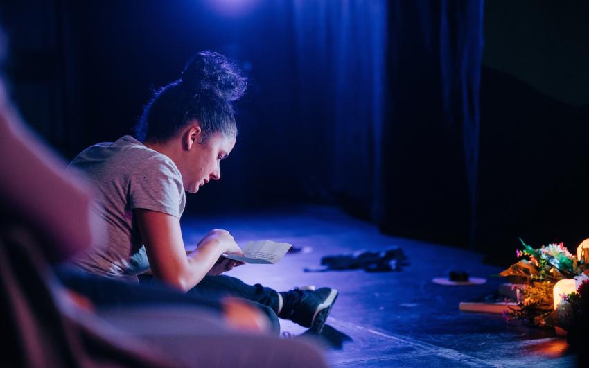 A performer on stage with her hair in a bun wearing a grey tshirt. She is reading a piece of paper.