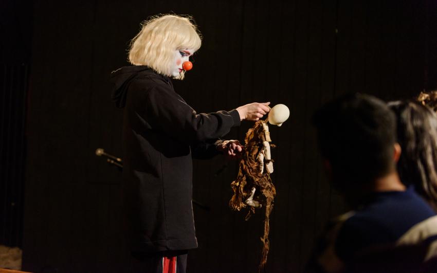 Jack wearing a blonde bob, white face paint, and a red nose, holding a puppet