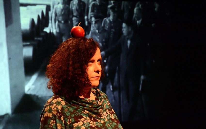  A woman standing rigidly with a red apple balanced on her head in front of a screen projecting historic images of Nazi soldiers