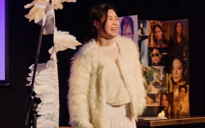 A landscape photo of a young East Asian woman in a white fur jacket laughing on a stage, next to a mic stand with feathers hanging down. Behind her are a cardboard shrine to Lucy Liu on a wooden table and flameless tea lights.