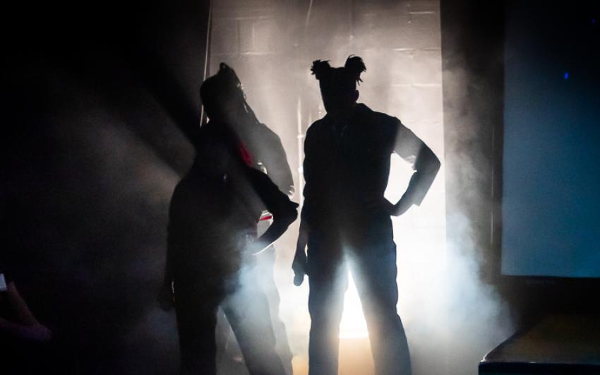 Three performers in silhouette on a dark stage with smoke billowing around them, lit up from beneath.