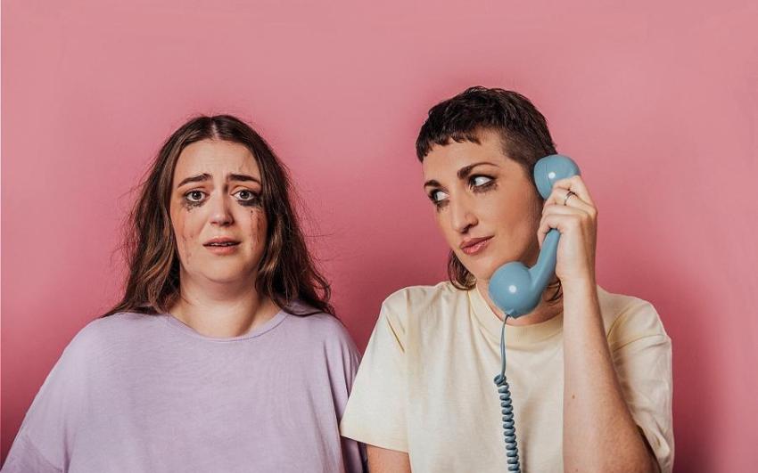 CARRIE RUDZINSKI and OLIVIA HALL in plain tshirts against a pink packground. They have mascara running down their cheeks as fi they've been crying and Carrie is holding a blue corded phone.