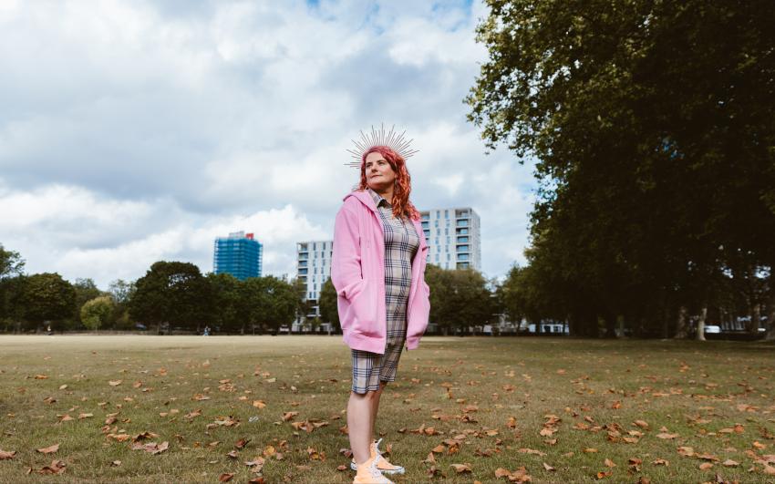 Kelly stands in a London park with three tower blocks in the background. She is wearing a large pink hoodie, a gold spikey crown, a check dress and yellow converses. it is an autumnal day with leaves on the ground.