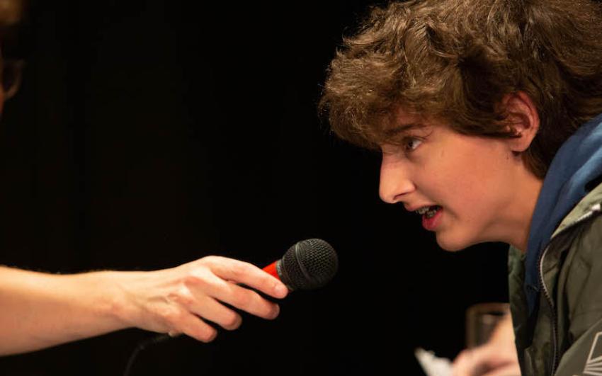 A young person with dark hair talks excitedly into a microphone.