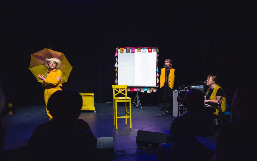Three performers on stage during a performance of ADHD The Musical. One is dancing with a yellow umbrella, one is singing into a standing microphone, one is sat playing keyboard and singing into a microphone.