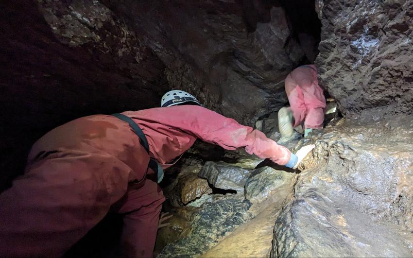 A person in a red caving suit clambering up the rock wall of a cave. Another person is disappearing through a crack in the rock ahead of them.
