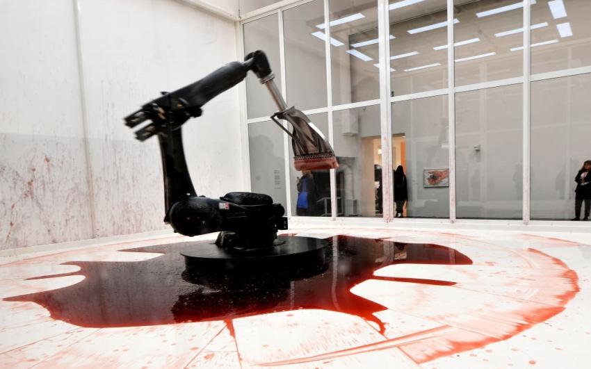 A display at the Guggenheim Gallery. A robot arm holding a squeegee stands in a pool of reddish-brown liquid in an otherwise white room. The room is streaked and splattered with the liquid. 