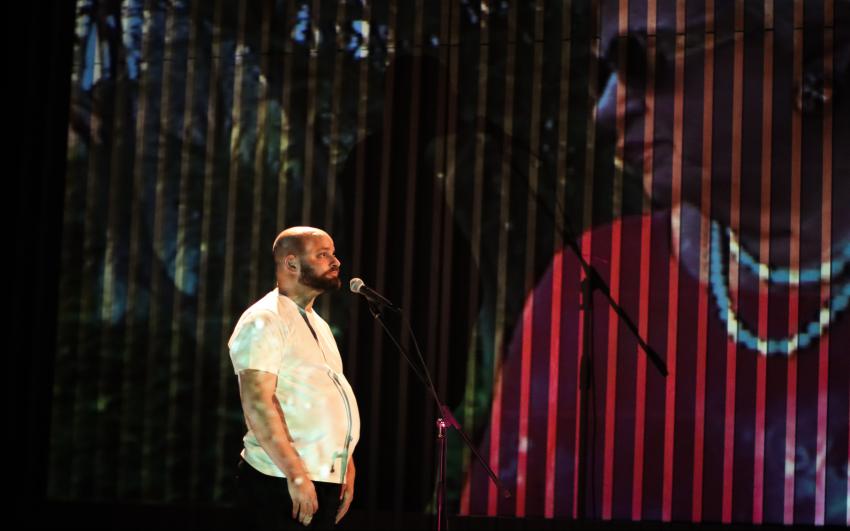 A man wearing in a white t-shirt stands centre stage, speaking into a microphone. He is looking up at a large projection of an older woman which takes up the entire back wall of the set.