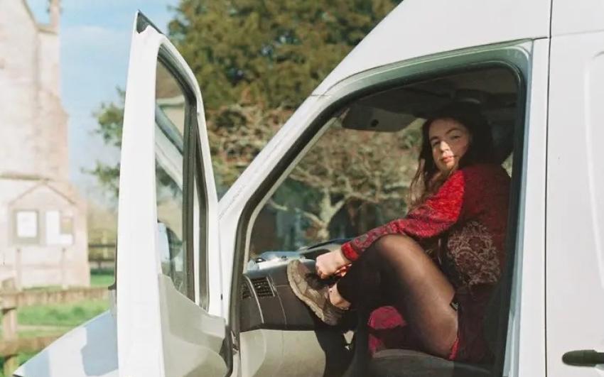 A white woman with long brown hair sits in the passenger seat of a white van. She is wearing a red patchwork jacket, black tights and trainers. The van door is open and she has her foot up on the dashboard tying her shoelace. 