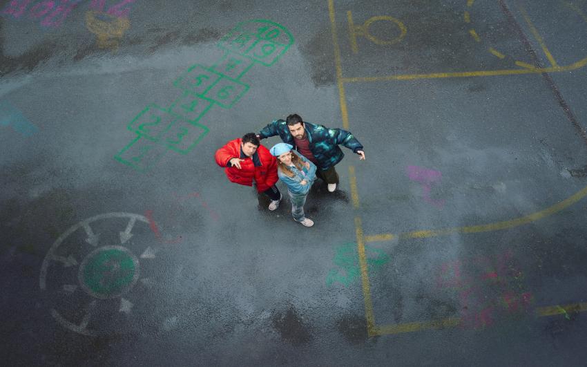 A woman and two men pose in a triangle formation in an empty school playground with various chalk markings on the ground.