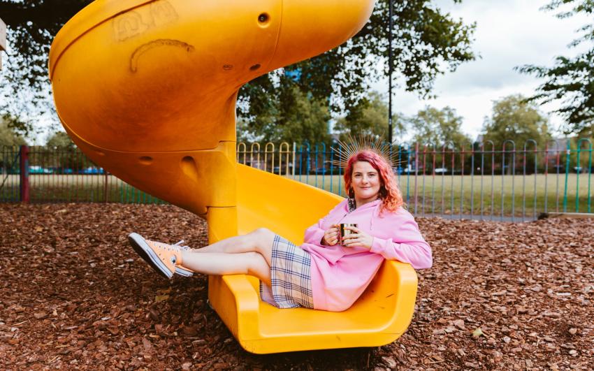 Kelly is a woman in 40s with pink and peach hair. She is wearing a halo crown, a pink hoodie over a checked dress with orange converse. She is sitting at the foot of a yellow slide in the park playground holding a gold mug. 