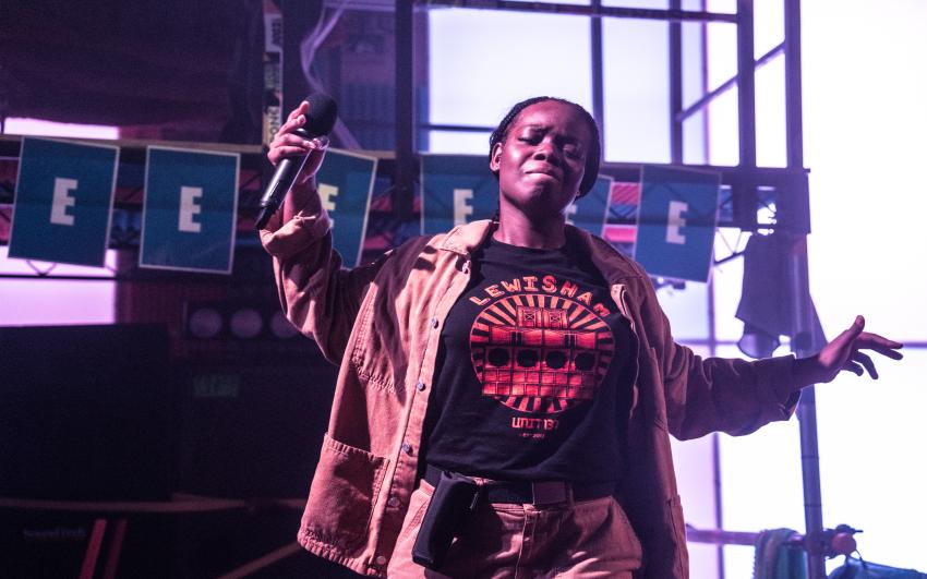 Zakiyyah Deen (a brown skinned black woman) holds a microphone in one hand and extends their other arm while performing. They are wearing a jacket over a t-shirt with a colorful graphic design.