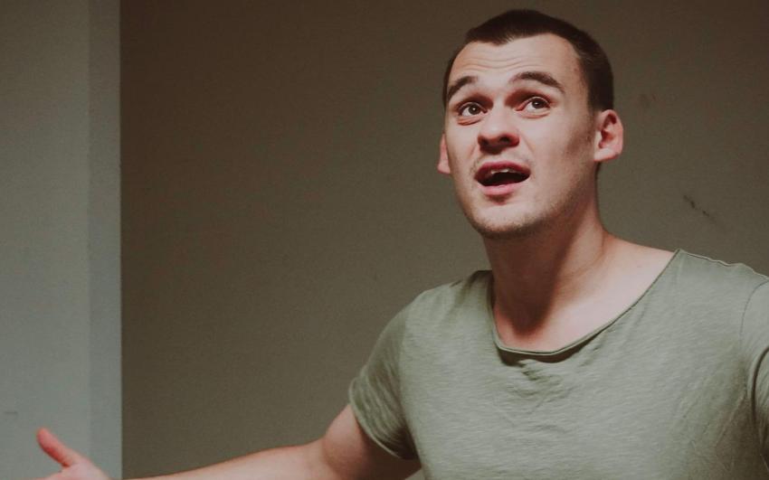 A white man with short dark hair wearing a green shirt, gestures with his arms out and mouth slightly open.